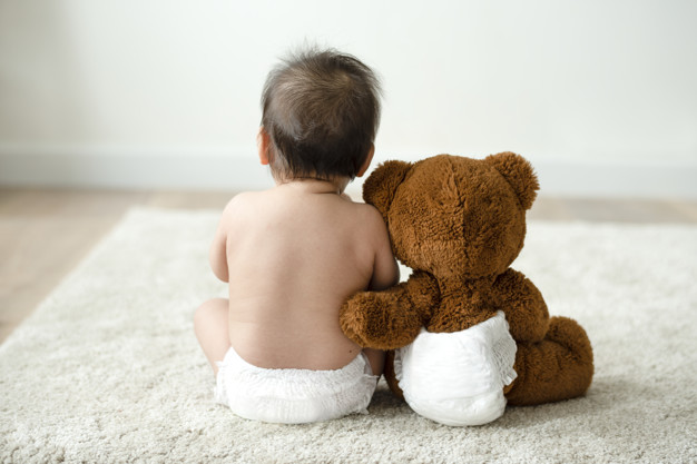 Seorang bayi sedang duduk bersama boneka beruang berwarna cokelat besar di sebelahnya sebagai simbol penyakit achondroplasia yang rentan dialami anak-anak