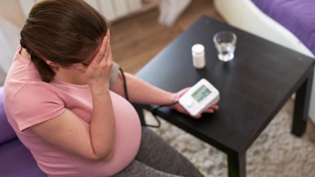 Seorang wanita hamil berbaju pink sedang mengukur tekanan darah untuk cek hipertensi secara mandiri di rumah